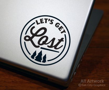 Photo of black vinyl decal of the phrase Let's Get Lost inside a circular shape with a pine tree forest underneath shown on laptop ©Salt City Graphics