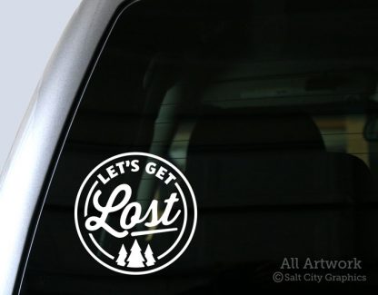 Photo of white vinyl decal of the phrase Let's Get Lost inside a circular shape with a pine tree forest underneath shown on truck window ©Salt City Graphics